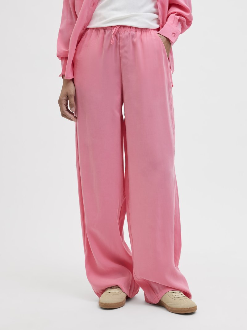 Personne portant un pantalon satiné rose ample, une chemise rose, un haut blanc et des baskets beiges, debout devant un fond clair uni.