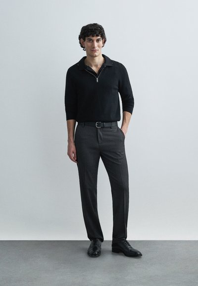 Black zip-neck sweater, gray tailored pants, and black shoes. Smooth fabric, fitted design, and discreet belt accent. Plain background.