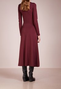 Sukně s dlouhými rukávy, v barvě maroon, vyrobená z hladkého materiálu, rozšiřující se do délky po kolena. Nosič nosí černé vysoké boty s chunky podpatkem.