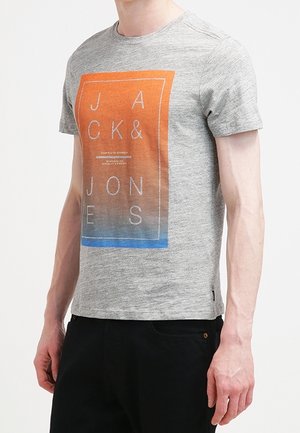 Mand iført en grå t-shirt med et rektangulært orange-til-blåt gradientprint, der viser teksten "JACK & JONES" med hvide bogstaver.