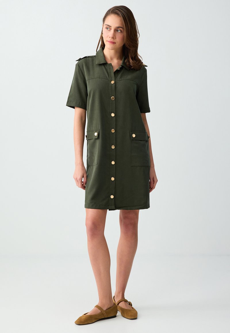 Femme portant une robe vert olive à manches courtes avec boutons sur le devant, poches à rabat et boutons dorés, associée à des chaussures plates marron.