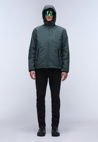 Chaqueta aislante verde oscuro con capucha, con cremallera frontal y dos bolsillos laterales, combinada con pantalones negros y gafas de sol reflectantes brillantes.
