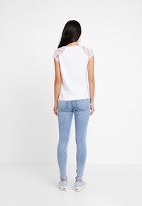 Bílá tričko s krajkovými krátkými rukávy, spárované s světle modrými skinny džíny a slip-on botami. Tkanina je hladká a střih je volný.