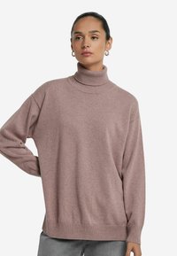 Kobieta o ciemnych włosach w niskim kokiem, nosząca złote kolczyki w kształcie kół, fioletowy sweter typu golf oraz szare spodnie, pozująca z jedną ręką na biodrze.