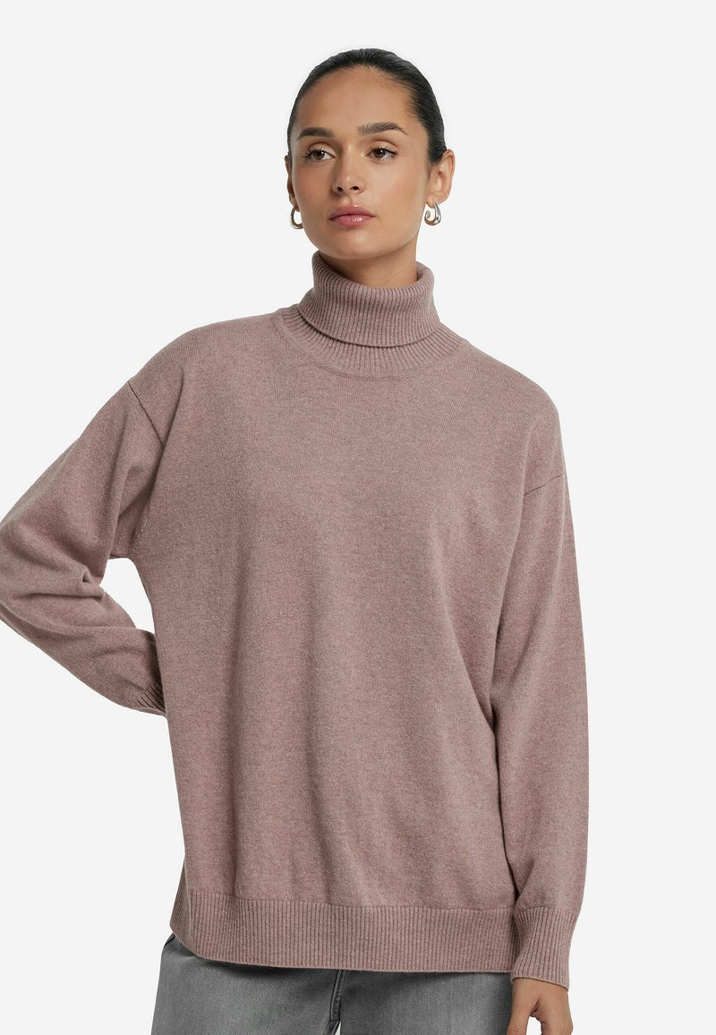 Kobieta o ciemnych włosach w niskim kokiem, nosząca złote kolczyki w kształcie kół, fioletowy sweter typu golf oraz szare spodnie, pozująca z jedną ręką na biodrze.