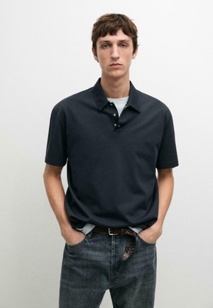 Jonge man met kort bruin haar die een donkerblauw poloshirt met korte mouwen draagt, ingestopt in verwassen spijkerbroek met een gevlochten riem, handen in de zakken.