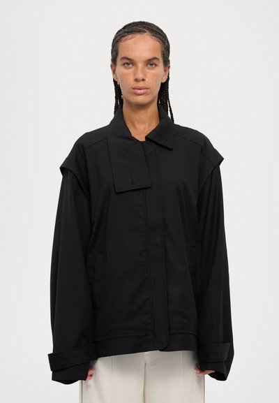Black oversized jacket with a large collar, side pockets, and wide sleeves. Made of smooth fabric, features subtle stitching details.