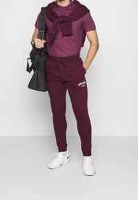 Man med bordeauxfärgad polo, matchande joggers med vit text, vita sneakers och bärande en svart väska med tennisrackethandtaget synligt.