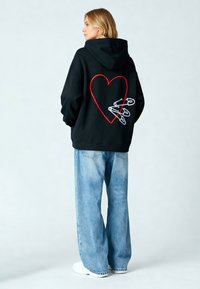 Felpa nera con cappuccio e design di un cuore rosso, con spille di sicurezza ricamate sul retro. Il modello indossa jeans larghi blu chiaro e scarpe bianche.