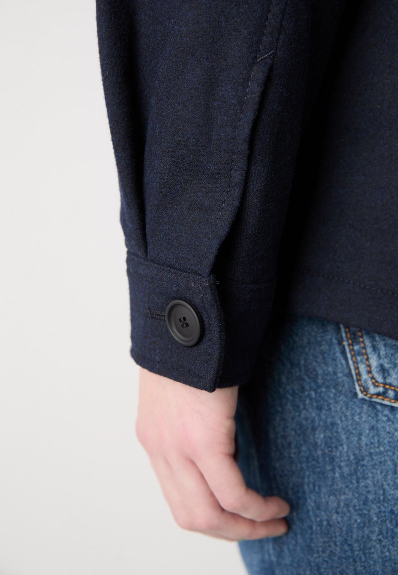 Chemise en laine bleu marine boutonnée avec un bouton noir et des détails de poignet cousus, assortie à un jean en denim clair.