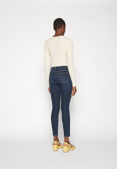 Jean skinny bleu foncé avec une taille haute, dotée de poches arrière, portée avec un haut crème à manches longues et des sandales à talons blocs jaunes.