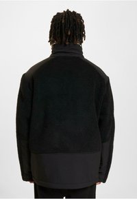 Schwarze Fleecejacke mit strukturierter Oberfläche, ausgestattet mit einem hohen Kragen und kontrastierendem schwarzem Stoff an Schultern und Ärmeln.