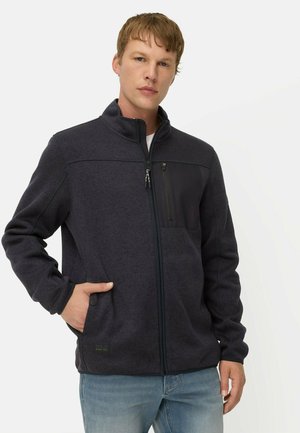Young man wearing a dark zip-up jacket with a chest zipper pocket and light blue jeans, standing against a plain light background.