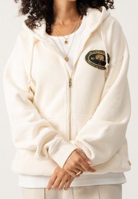 Roomkleurige hoodie met rits van zachte stof, met een geborduurd groen- en goudkleurig logo op de borst, verstelbare trekkoorden en oversized mouwen.