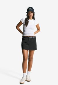 White crew-neck t-shirt, black denim skirt with belt, white athletic socks, beige sneakers. Baseball cap with text, straight posture.