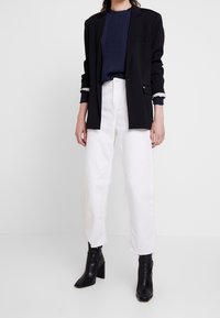 Blazer noir sur mesure, top plissé bleu marine, pantalon blanc raccourci et bottines noires à talons carrés. Lignes  épurées et couleurs contrastées.