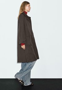 Langer, dunkelbrauner Mantel mit weiten Ärmeln und Knopfverschlüssen, kombiniert mit einem roten Rollkragenpullover und hellblauen weiten Jeans. Schwarze Sandalen.