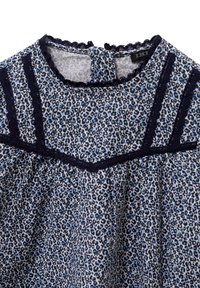 Top in tessuto leopardato blu e bianco con rifiniture blu navy, dotato di bordi scallop, scollatura rotonda e bande verticali decorative.