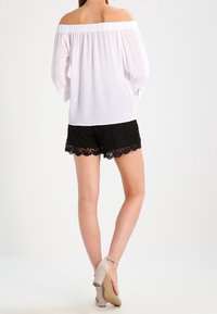 Biała bluzka bez ramiączek z elastycznym wykończeniem, w połączeniu z czarnymi szortami wykończonymi koronką. Modelka nosi beżowe sandały na obcasie.
