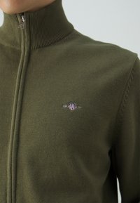 Olivgrüner Pulli mit hohem Kragen, Reißverschluss vorne und glatter Textur. Verfügt über ein kleines Logo auf der Brust.