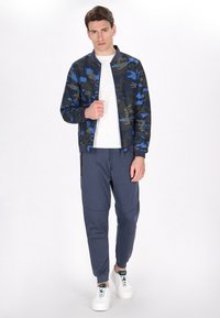 Giacca bomber camouflage in tonalità blu e verde, abbinata a pantaloni jogger blu navy. Una maglietta bianca e sneaker bianche completano l'outfit.