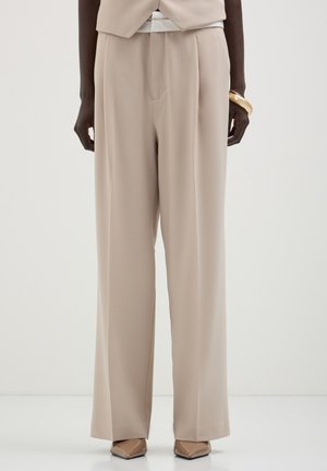 Pantalons larges beige en tissu doux, dotés d'une taille mi-haute, de plis avant et d'une texture lisse. Coupe classique avec un minimum de détails.