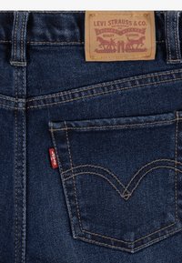 Tmavě modré džíny z denimu s klasickým pětikapsovým designem. Obsahují koženou nášivku a červenou logo stuhu na zadní kapse. Kontrastní šití.