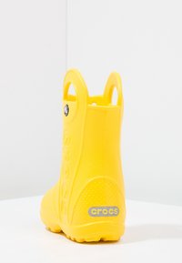 Ljusgul Crocs-regnboot med draghandtag och texturerad sula, visad bakifrån på en vit yta och ljus bakgrund.
