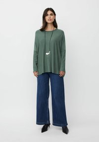 Locker sitzender, langärmeliger grüner Pullover kombiniert mit weit geschnittenen, dunkelblauen Jeans. Eine schlichte silberne Anhänger-Halskette rundet das Outfit ab.
