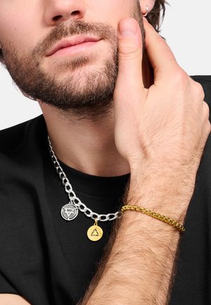 Bärtige Person, die ein schwarzes Hemd trägt und ihr Gesicht berührt, mit sichtbarer silberner Halskette und goldenem Kettenarmband.