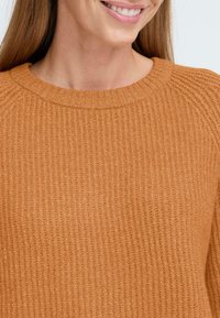 Megztinis šiltos gintarinės oranžinės spalvos. Yra raukšlėta tekstūra, apvalus iškirpimas ir subtilūs siuvinėjimo akcentai pečių srityje.