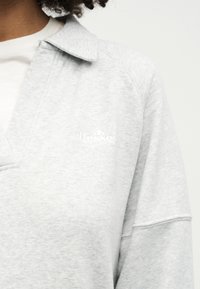 Άτομο που φοράει ανοιχτό γκρι φούτερ με γιακά και μικρό λευκό λογότυπο ellesse στο αριστερό στήθος, με σπαστά μαλλιά ορατά στον λαιμό.