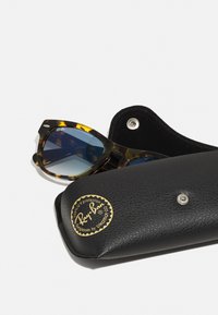 Óculos de sol de tartaruga castanha com lentes azuis dentro de uma capa de pele preta com um logótipo dourado e fecho de pressão.