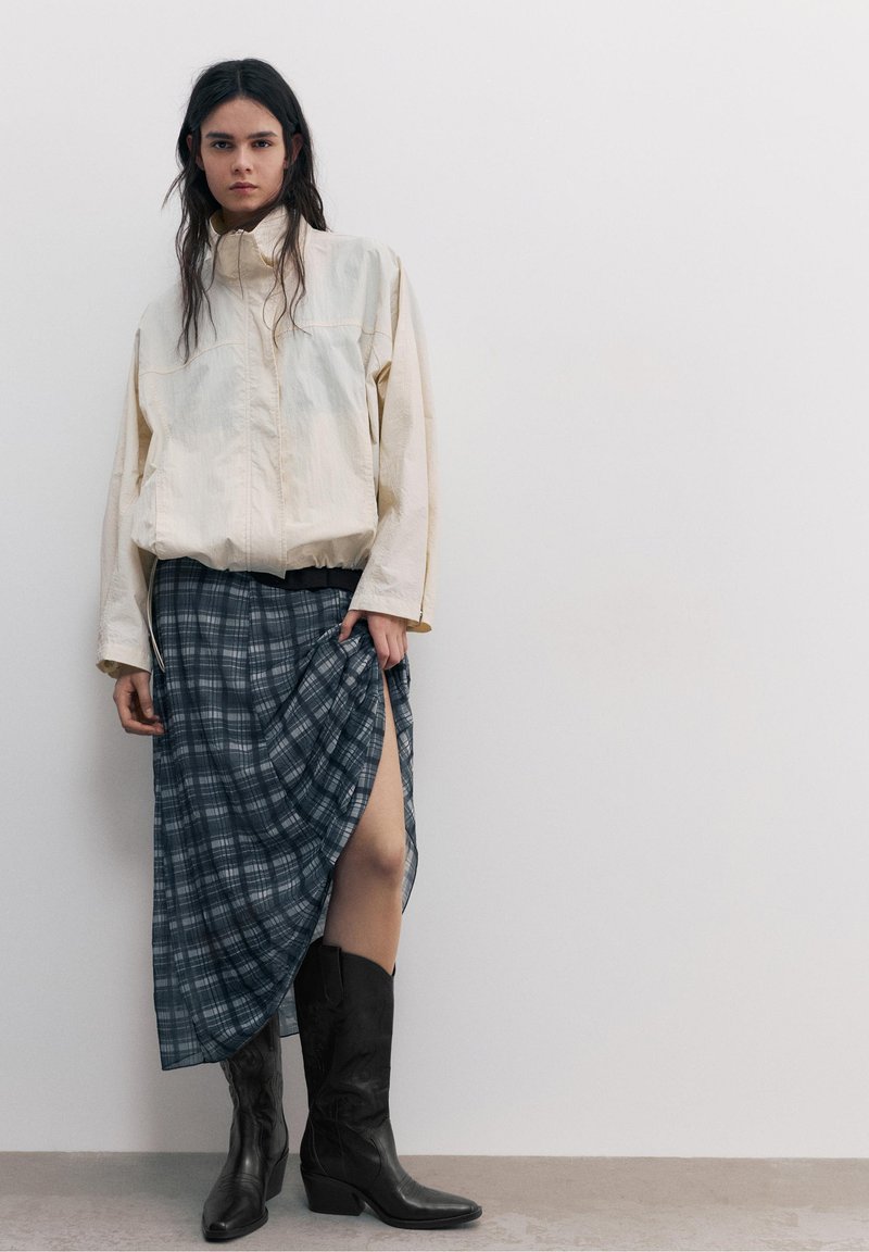 Tânără cu păr lung și închis la culoare poartă o jachetă ușoară, fustă în carouri cu o despicătură pe coapsă și cizme negre până la jumătatea gambei, stând în fața unui perete simplu.