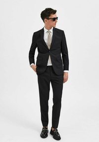 Svart dress med slim fit, med en enkelspontet jakke, hvit skjorte og teksturert beige slips. Svart loafers med gullaksenter.