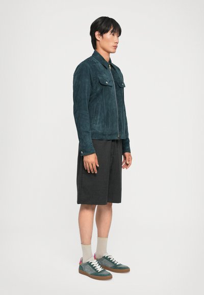 Veste en daim bleu avec fermeture zippée, poches avant, associée à un short noir et des baskets vertes avec des accents colorés et des lacets blancs.