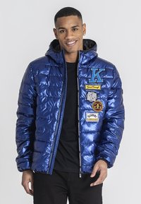 Blå pufferjacka i glänsande material, med huva, dragkedja och flera lappar inklusive bokstäver och logotyper på framsidan.