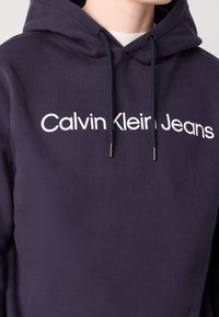 Marineblå hoodie av mjukt tyg, med en framtida logotyp "Calvin Klein Jeans" i stora vita bokstäver, samt dragsnören för huvan.