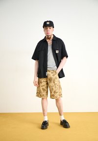 Black short-sleeve shirt over gray tank top, paired with tan camo shorts. Black cap and footwear complete the look. White socks visible.