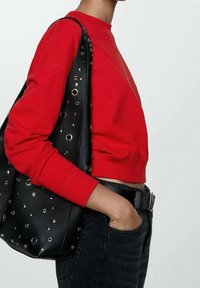 Person trägt einen roten Cropped-Sweatshirt und schwarze Jeans und trägt eine schwarze Ledertasche mit silbernen Ösen über der Schulter.