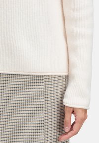 Jasnobeżowy sweter w prążki z długimi rękawami, w połączeniu z kraciastą spódnicą w czarno-brązowo-kremowym wzorze.