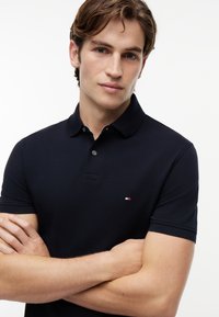 Young man with light brown hair wearing a black short-sleeve polo shirt, arms crossed, neutral expression on white background.