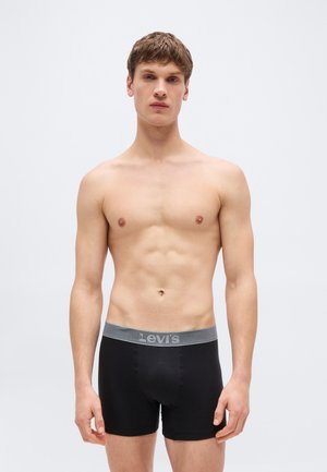 Jeune homme aux cheveux bruns courts debout, portant un boxer Levi's noir avec une ceinture grise, sur un fond clair uni.