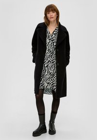 Schwarzer, flauschiger Mantel mit Revers und Knöpfen, getragen über einem kurzen weißen Kleid mit schwarzem Zebra-Muster, kombiniert mit schwarzen klobigen Stiefeln.
