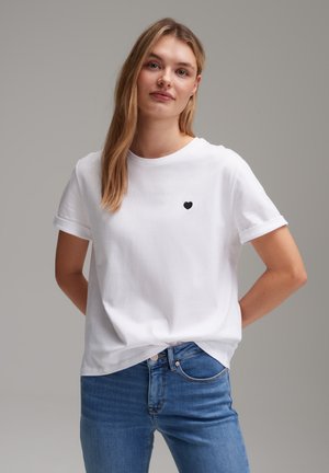 T-shirt en coton blanc à manches courtes retroussées, avec un petit emblème de cœur noir sur la poitrine. Associé à un jean bleu.