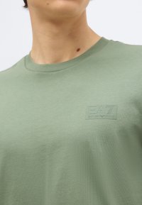 Person som har på seg en lysegrønn skjorte med rund hals og preget EA7 Emporio Armani-logo på venstre bryst.