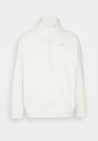 Hvid Nike pullover-jakke med høj krave, halv-zipped front, elastiske ærmer og kant; har et diskret chevronmønster og logo.