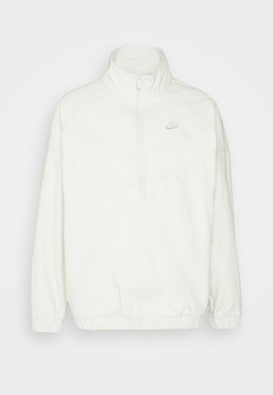 Jachetă albă Nike cu guler înalt, fermoar pe jumătate în față, manșete elastice și tiv; prezintă un model subtil în formă de V și logo.