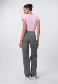 Frau mit einem Dutt, die ein pinkes kurzärmliges Oberteil, hoch geschnittene graue Jeans und weiße Sneakers trägt, steht mit dem Rücken zu mir.