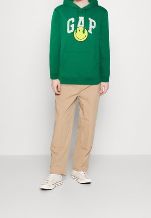 Sweat à capuche vert avec le logo "GAP" et un graphique smiley jaune, assorti à un pantalon cargo beige et des baskets blanches. Tissu et coupe décontractés.
